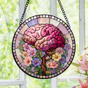 Op de afbeelding: Een ronde glas-in-lood zonnevanger met een roze en paars menselijk brein in de vorm van een boom, met een stam die uitloopt in een cluster van kleurrijke bloemen. Het stuk is omlijst door een decoratieve rand en hangt aan een zwarte ketting.