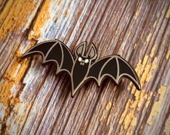 May include: Black enamel bat pin with white eyes. The pin is on a wood background.
