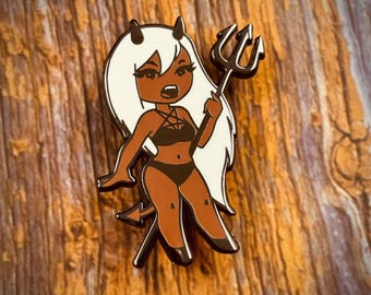 May include: A black and white enamel pin featuring a cartoon character with white hair, horns, and a black bikini holding a trident. The character has brown skin and a black choker with a cross.