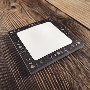 May include: A stack of square notepads with a black border featuring a moon phase design. The centre of the notepad is white, providing space for notes. The notepads are on a wooden surface.