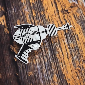 May include: A silver and black toy ray gun pin. The pin has a detailed design with a black handle and a silver barrel. The pin is on a wooden surface.