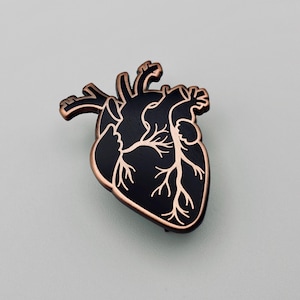 May include: A black anatomical heart-shaped pin with copper-colored outlining and veins. The pin is a detailed representation of a human heart, suitable for medical professionals or those interested in anatomy.