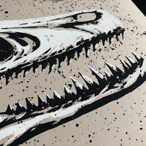 May include: Close-up of a black and white illustration of a dinosaur skull. The artwork features sharp teeth and a detailed bone structure against a beige background with black speckles. The image is a print.