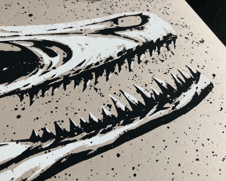 May include: Close-up of a black and white illustration of a dinosaur skull. The artwork features sharp teeth and a detailed bone structure against a beige background with black speckles. The image is a print.