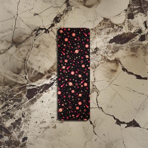 Pode incluir: Um marcador de página preto com um padrão repetido de planetas, estrelas e estrelas cadentes vermelhas e laranja.