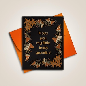 Op de afbeelding: Zwarte wenskaart met de tekst "I love you my little trash gremlin!" in goud, omringd door oranje bloemen en paddenstoelen. Inclusief een oranje envelop.