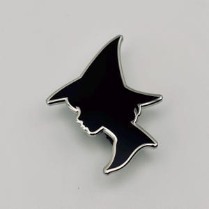 May include: A black enamel witch silhouette pin with a silver-colored border. The pin features the profile of a witch wearing a pointed hat. The pin is a decorative accessory.