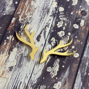 May include: Two gold-coloured metal deer antlers, each with three points, are shown against a rustic wooden background.