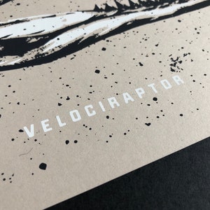May include: Close-up of a print featuring a stylized dinosaur illustration. The artwork is in black and white, with the word "VELOCIRAPTOR" printed in white block letters. The background is a speckled tan color.