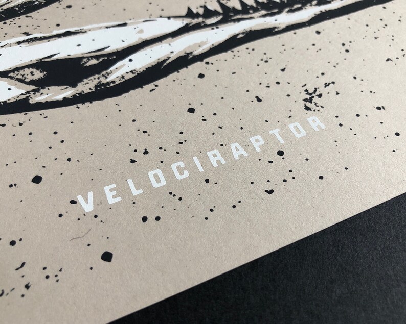 May include: Close-up of a print featuring a stylized dinosaur illustration. The artwork is in black and white, with the word "VELOCIRAPTOR" printed in white block letters. The background is a speckled tan color.