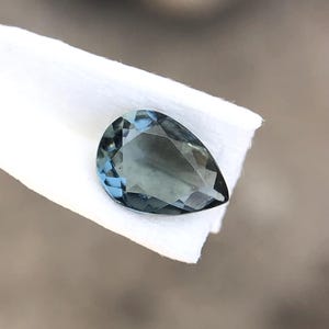 Könnte beinhalten: Ein tropfenförmiger, facettierter Edelstein in Blau- und Türkistönen. Der Stein liegt auf einer weißen Oberfläche, die seine Klarheit und seinen Schliff hervorhebt. Die Facetten des Edelsteins reflektieren das Licht.