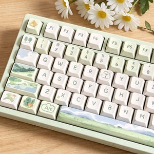 May include: A mint green mechanical keyboard with custom keycaps featuring landscape and nature-themed designs. The keyboard has white keycaps with illustrations of trees, mountains, and clouds. White daisies with yellow centres are in the background.