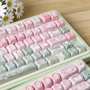 Może przedstawiać: Mechaniczna klawiatura w pastelowych kolorach z różowymi, zielonymi i białymi nakładkami na klawisze. Nakładki na klawisze mają kwiatowe i wzorzyste wzory. Bukiet stokrotek spoczywa obok klawiatury. Klawiatura ma jasnozieloną ramkę.