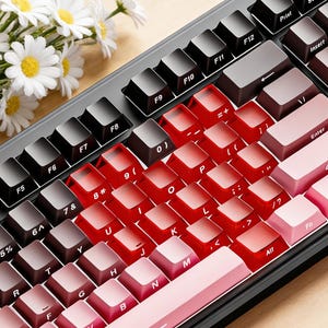 May include: A mechanical keyboard with black, red, and pink keycaps. The keys are arranged in a standard layout, with a gradient effect on the red keys. A bouquet of white daisies rests on the wooden surface.