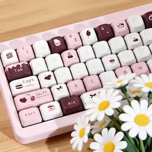 May include: A pink mechanical keyboard with custom keycaps featuring dessert and heart designs. The keyboard has white, pink, and brown keys. Daisies are in the foreground.