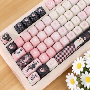 May include: A custom mechanical keyboard in pink and white. The keycaps are decorated with cat paw prints, cat illustrations, and a checkered design. The keyboard has a light pink frame and keys in black, white, and pink.