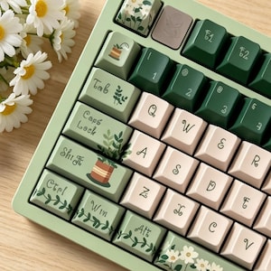 May include: A mechanical keyboard with a light green frame and custom keycaps featuring botanical illustrations of potted plants, vines, and white daisies. The keys are a mix of cream, dark green, and light green, with stylized lettering.
