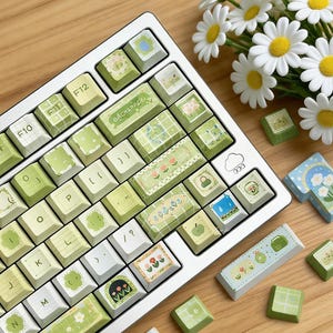 Può includere: Una tastiera bianca con tasti personalizzati nei toni del verde e del bianco, con vari disegni a tema naturale. I tasti presentano illustrazioni di fiori, nuvole e altri elementi decorativi. Un mazzo di margherite è nelle vicinanze.