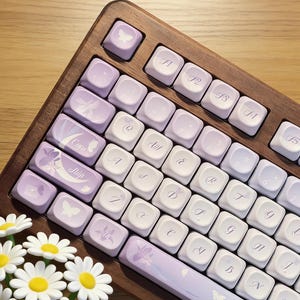 Puede incluir: Un teclado personalizado con marco de madera y teclas blancas y lavanda. Las teclas presentan diseños florales y de mariposas. El teclado está sobre una superficie de madera, con un ramo de margaritas en primer plano.