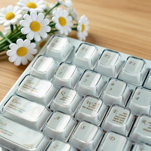 May include: A close-up of a white keyboard with clear keycaps. The keycaps have gold lettering and symbols. A bouquet of white daisies with yellow centers is in the background.