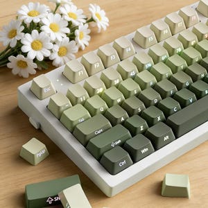May include: A mechanical keyboard with a gradient green and white keycap set. The keys are arranged in a standard layout with alphanumeric keys, function keys, and modifier keys. The keyboard is on a white base. A bouquet of white flowers is in the background.