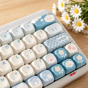 May include: A computer keyboard featuring a custom keycap set in shades of white and light blue. The keys display various icons including a cloud, a paw print, flowers, and a small animal face. The enter key has a blue and white checkered pattern.