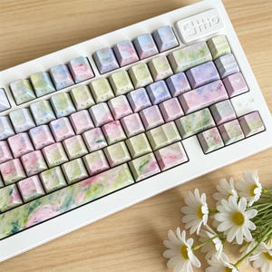 May include: A white mechanical keyboard featuring custom keycaps with a soft, watercolor-style pattern in shades of pastel pink, light green, and lavender. The keys display standard alphanumeric characters and function labels in a dark font.