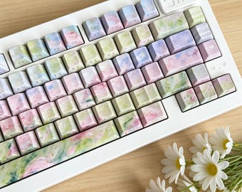 May include: A white mechanical keyboard featuring custom keycaps with a soft, watercolor-style pattern in shades of pastel pink, light green, and lavender. The keys display standard alphanumeric characters and function labels in a dark font.