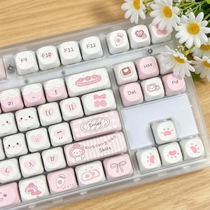Puede incluir: Un teclado mecánico con teclas blancas y rosa pastel decoradas con varias ilustraciones que incluyen corazones, cerezas, lazos, huellas de patas y personajes. Las teclas muestran texto como Enter, Shift, Del y de F9 a F12.