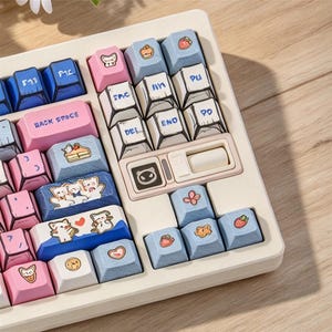 May include: A mechanical keyboard with a cream-coloured frame featuring custom keycaps in shades of blue, pink, and white. The keys display various illustrations including cats, strawberries, and bread, alongside text labels like BACK SPACE, DEL, and END.