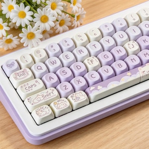 May include: A mechanical keyboard with a white frame and a mix of light purple and cream-colored rounded keycaps. The keys feature purple lettering, with specific keys displaying illustrations of cats, a bow, and clouds. The spacebar has a cloud design.