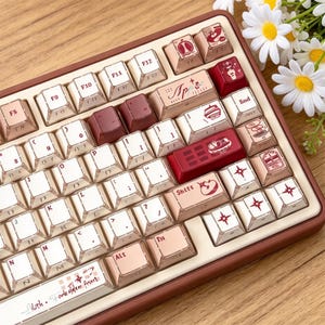 May include: A mechanical keyboard with a brown frame and a mix of white, light pink, and dark red keycaps. Several keys feature custom graphic icons, including stars, food items, and text such as App, Shift, and Lilith Forbidden Fruit.