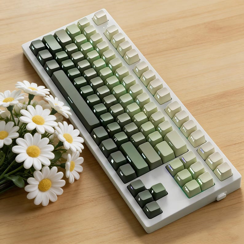 May include: A white mechanical keyboard with green gradient keycaps. The keyboard is on a wooden surface, next to a bouquet of white daisies with yellow centers.