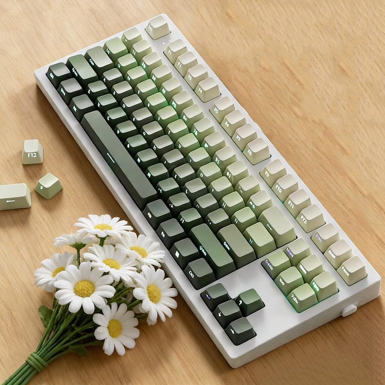 May include: A white mechanical keyboard with green gradient keycaps. The keyboard has a variety of keys, including function keys and a space bar. A small bouquet of white daisies with yellow centers rests near the keyboard.