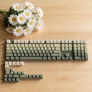 May include: A mechanical keyboard with green and off-white keycaps is displayed on a wooden surface. The keyboard is partially disassembled, with some keycaps removed. A bouquet of white daisies with yellow centers is in the background.