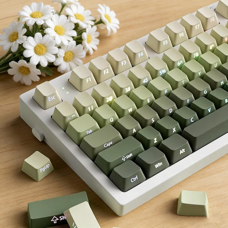 May include: A mechanical keyboard with a green and white gradient keycap set. The keys are arranged in a standard layout with alphanumeric, function, and modifier keys. The keyboard sits on a white base. A bouquet of white flowers is in the background.