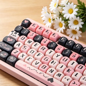 Puede incluir: Un teclado mecánico con teclas redondeadas en tonos rosa pastel y negro. Las teclas muestran caracteres alfanuméricos y símbolos en blanco. La barra espaciadora es rosa con un pequeño diseño de corazón pixelado. El teclado está colocado junto a un ramo de margaritas blancas.