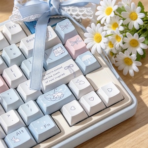 May include: A mechanical keyboard with light blue, white, and pale pink keycaps featuring icons of umbrellas, bows, and hearts. One keycap displays the text, "the storm passed but the scent of rain lingered." A light blue ribbon and white daisies are placed on top.