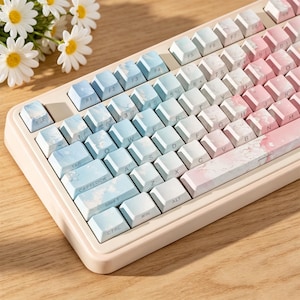 Peut inclure: Un clavier mécanique présentant un dégradé de couleurs passant du bleu clair à gauche au rose tendre à droite. Les touches affichent un motif marbré et le cadre du clavier est d'une couleur beige clair unie.