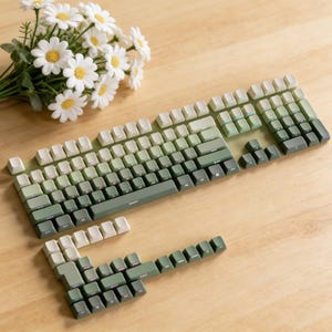 May include: A mechanical keyboard with a gradient color scheme, transitioning from white to various shades of green. The keyboard is disassembled, with keycaps scattered. A bouquet of white daisies with yellow centers rests nearby on a wooden surface.