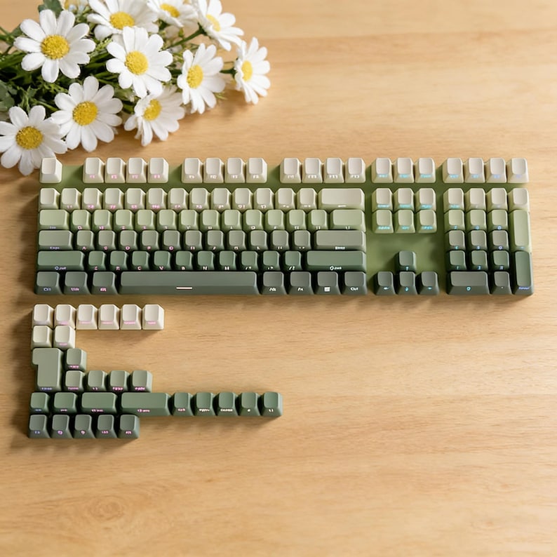 May include: A green and cream-colored mechanical keyboard with keycaps in varying shades of green. The keyboard is partially disassembled, with some keycaps removed. A bouquet of white daisies with yellow centers sits nearby.