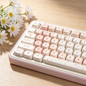 May include: A mechanical keyboard featuring a light pink frame and a color-blocked keycap set in shades of cream and soft pink. The keys display a standard QWERTY layout with brown lettering, and the spacebar is decorated with small pink petal accents.