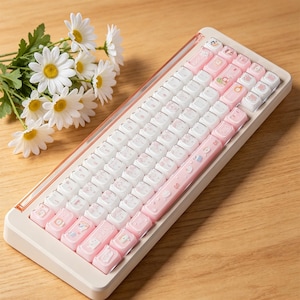 Puede incluir: Un teclado mec&aacute;nico con marco color crema y una mezcla de teclas blancas y rosa pastel. Las teclas presentan peque&ntilde;as ilustraciones gr&aacute;ficas coloridas de varios objetos. Un ramo de margaritas blancas con centros amarillos se encuentra al lado.