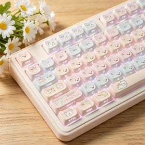 Puede incluir: Un teclado mecánico con marco beige claro y teclas de colores pastel con forma de orejas de gato e ilustraciones variadas que incluyen cerezas, tostadas y animales. Las teclas muestran letras, números y etiquetas de función.