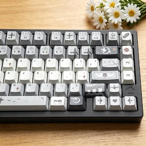 May include: A mechanical keyboard with custom keycaps in white, grey, and black. The keycaps feature various symbols and icons. The keyboard has a black frame and is on a wooden surface. A bouquet of white daisies is in the background.