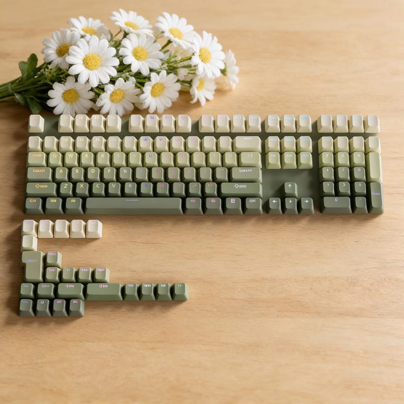May include: A green and cream-colored mechanical keyboard with keycaps in varying shades of green. A bouquet of white daisies with yellow centers rests beside the keyboard on a wooden surface. The keyboard is partially disassembled.