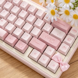 Puede incluir: Un teclado mecánico con teclas de color rosa claro que presentan varias ilustraciones y símbolos de gatos. La tecla enter muestra un gráfico de un gato y flores, mientras que un pequeño lazo de satén rosa está sujeto a la esquina inferior derecha.