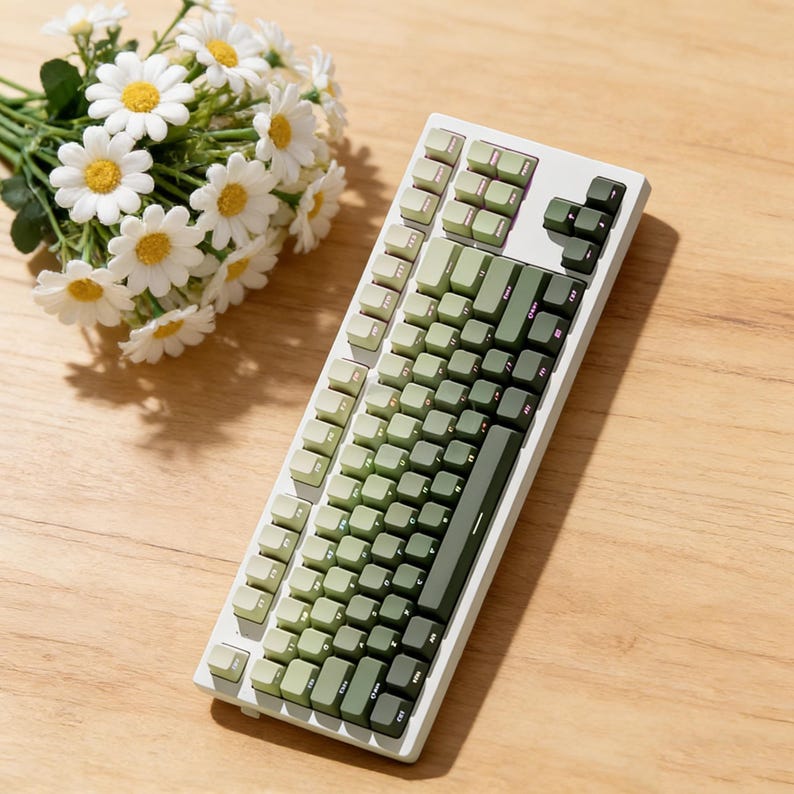 May include: A white mechanical keyboard with green keycaps in varying shades. The keyboard is angled, showing the keys in detail. A bouquet of white daisies with yellow centers sits to the left of the keyboard on a wooden surface.