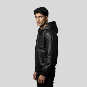 Peut inclure: Veste à capuche en cuir noir avec fermeture éclair. La veste a un ourlet et des poignets côtelés. Le modèle porte un pantalon noir. Le fond est gris clair uni.