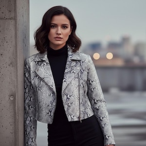 May include: A gray and white snakeskin-patterned motorcycle jacket with a black turtleneck and black pants. The jacket has a zipper closure and a classic biker jacket design. The model is leaning against a concrete pillar.